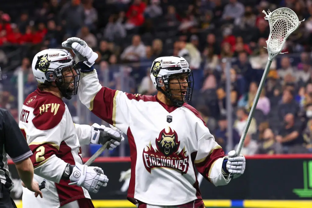 Two lacrosse players celebrating a goal on the field, wearing white and maroon jerseys with "Albany Firewolves" logo. One player raises his stick in joy, while the other smiles and raises his arm in a congratulatory gesture. Spectators are visible in the background.