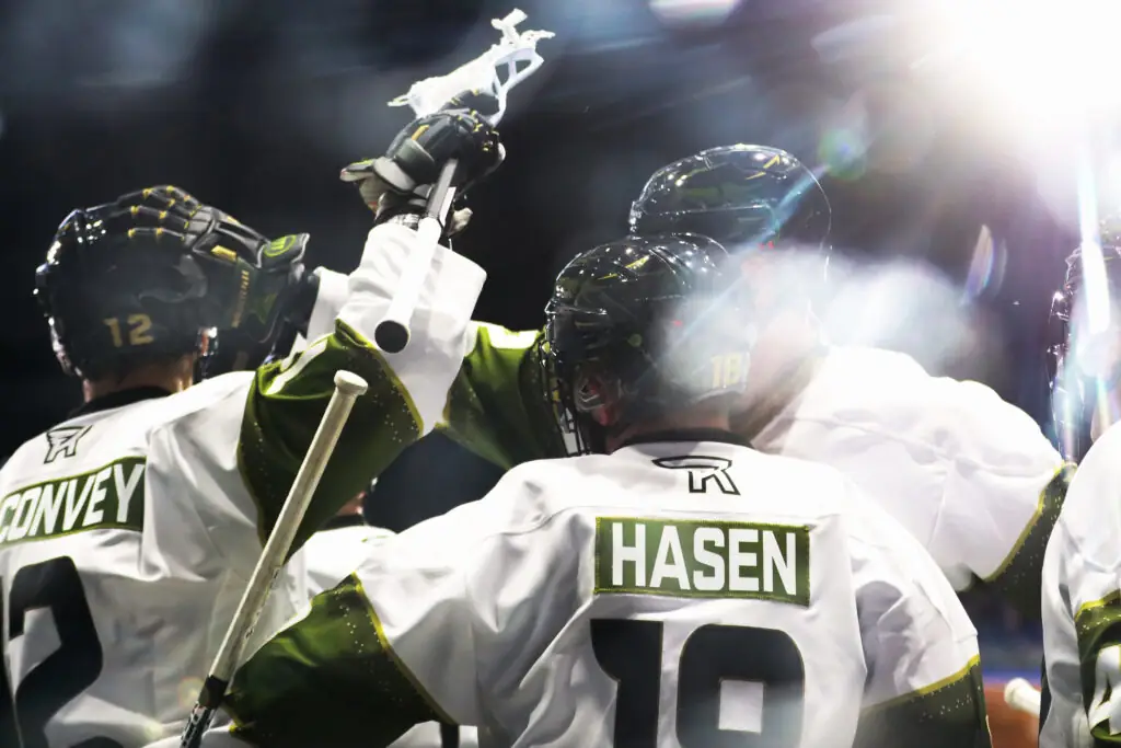 A group of lacrosse players celebrate, one player raises a lacrosse stick in the air. The jerseys are predominantly white with green accents, and player names like "HASEN" and "CONVEY" are visible on the backs.