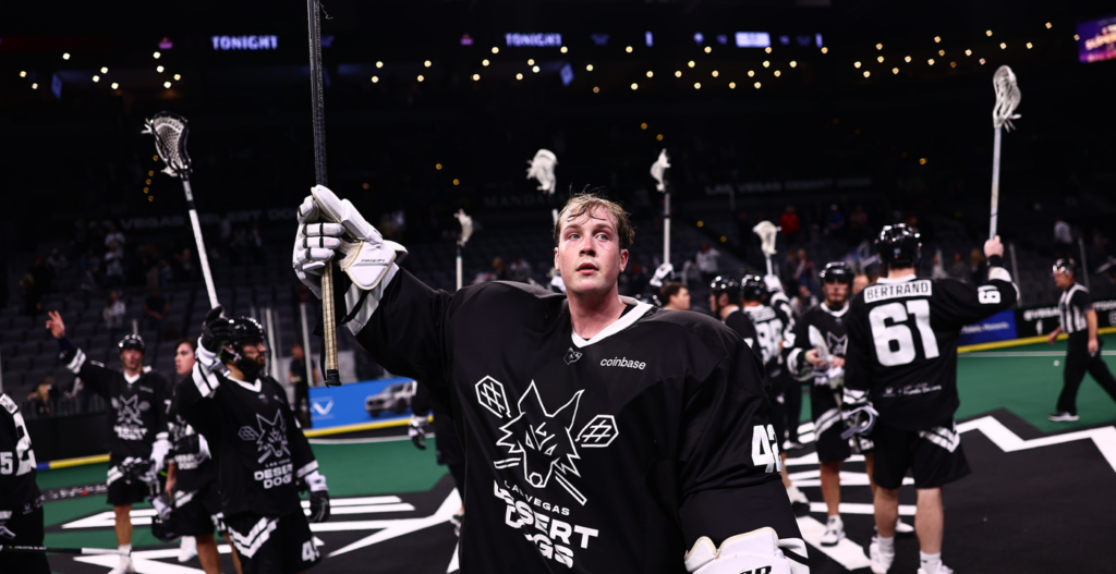 A lacrosse player wearing a black jersey and gloves holds a stick raised in celebration, with teammates in black jerseys celebrating in the background on a green field.