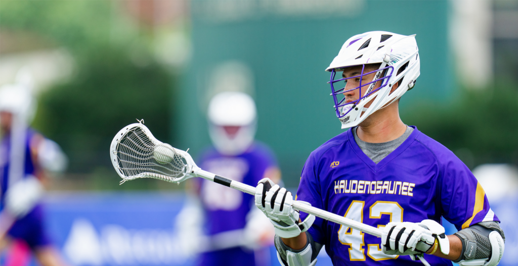 A lacrosse player in a purple jersey with "HAUDENOSAUNEE" and the number "43" stands holding a lacrosse stick with a net, focused on the game. Background features blurred teammates in jerseys.
