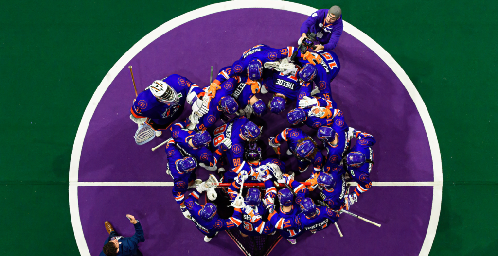 Aerial view of a group of ice hockey players in purple and orange uniforms huddled together on the ice, with a coach or staff member standing nearby.