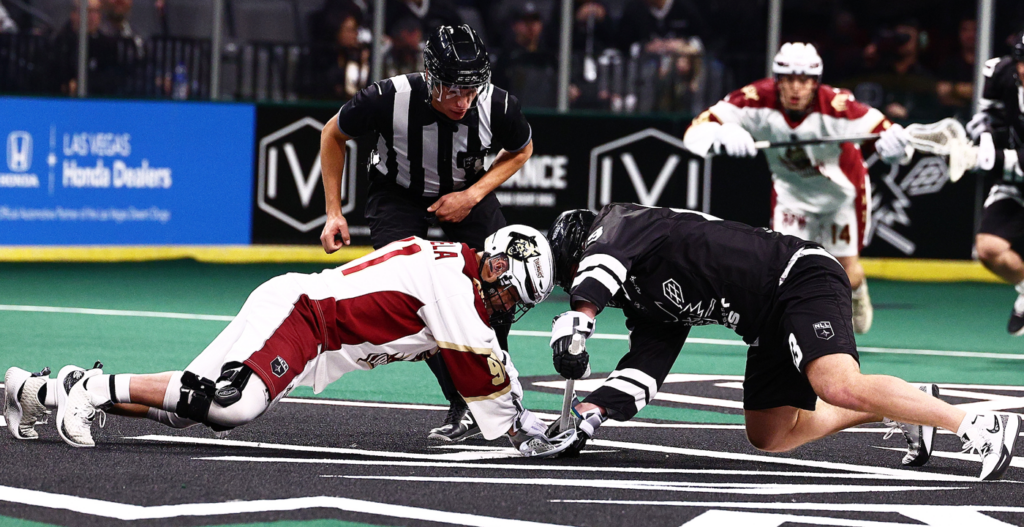 Two lacrosse players are facing off on the field, with one in a white and maroon uniform and the other in a black uniform. A referee is observing the faceoff, while other players are positioned in the background. The playing surface is green, and the surroundings are filled with spectators.