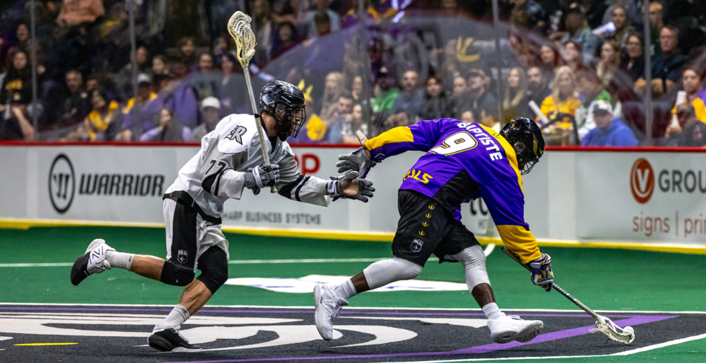 A lacrosse player in a white uniform is attempting to tackle another player in a purple and yellow uniform during a game. The scene shows intense action on the field, with spectators visible in the background.
