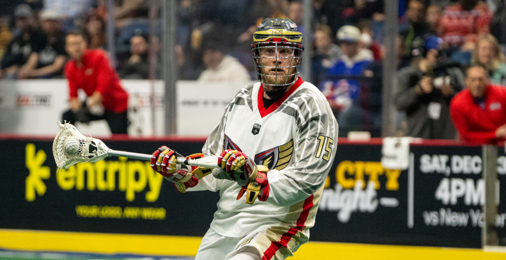 A lacrosse player wearing a grey and red jersey with the number 15 holds a lacrosse stick in a game setting, with a crowd visible in the background.