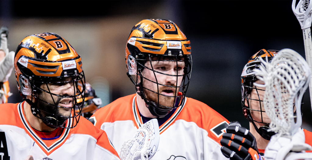 A group of lacrosse players wearing orange and black helmets and jerseys are engaged in a discussion on the field, with one player holding a lacrosse stick and showing intense expressions.