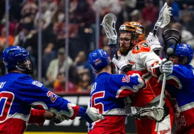 A lacrosse player wearing an orange and white uniform is being held and challenged by two players in blue and red uniforms during a game, with a crowd visible in the background.