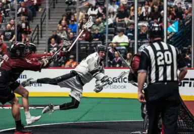 A lacrosse player in a white jersey is airborne attempting a shot, while two opponents in maroon jerseys defend against him. The scene captures the intensity of the game with spectators visible in the background and a referee in a black and white striped shirt nearby.