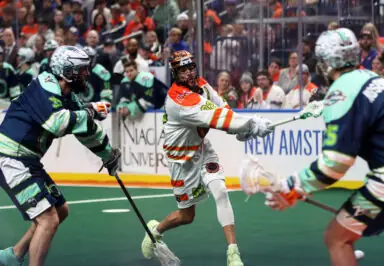 A lacrosse player in a white and orange uniform is in mid-action, passing the ball while being guarded by two players in dark blue and green uniforms. The scene captures the intensity of a lacrosse game, with a crowd visible in the background.