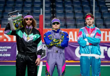 Three men dressed in colorful, retro athletic outfits pose together on a sports field. One man holds a boombox and wears sunglasses and a gold chain, while the others have their arms crossed and wear vibrant tracksuits with geometric patterns.