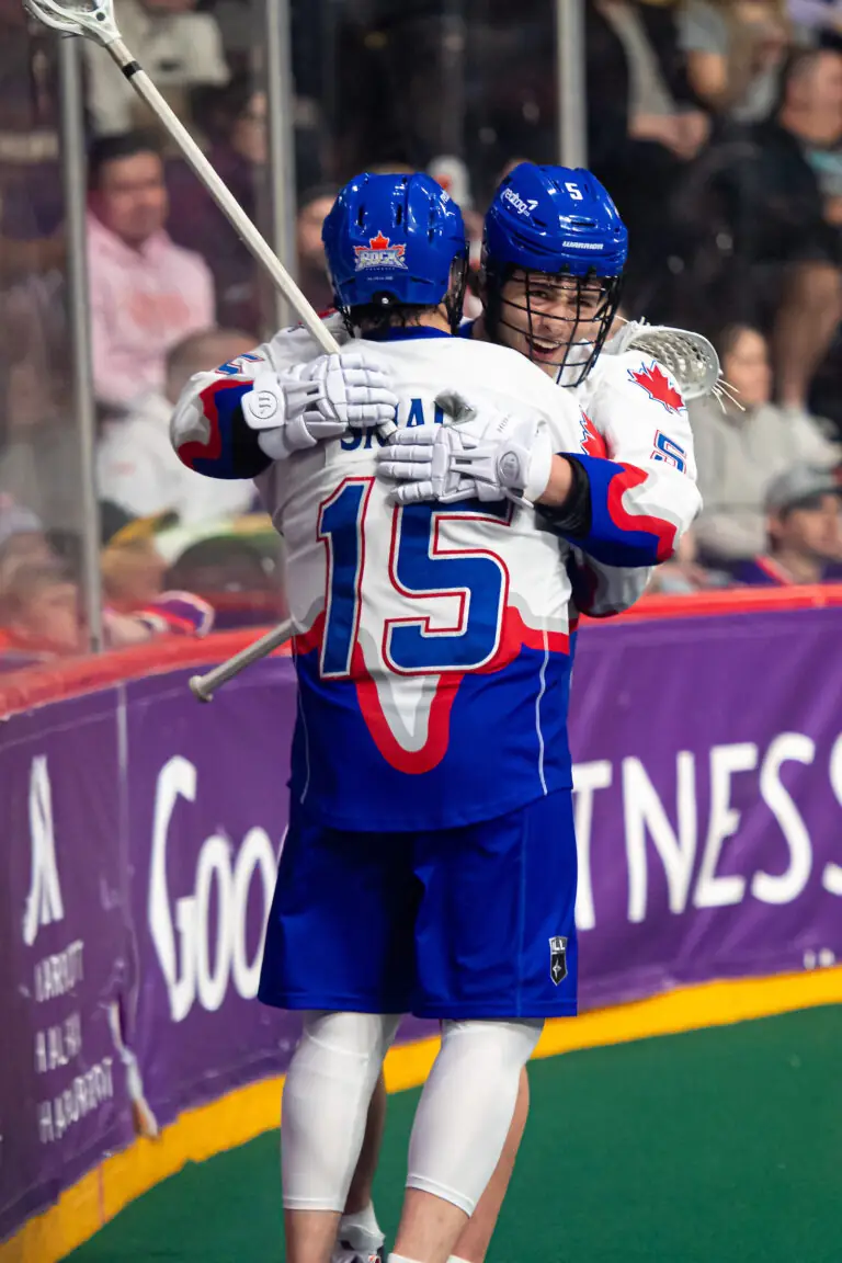 Two lacrosse players in blue and white jerseys embrace in celebration, with one wearing number 15. A cheering crowd is visible in the background.
