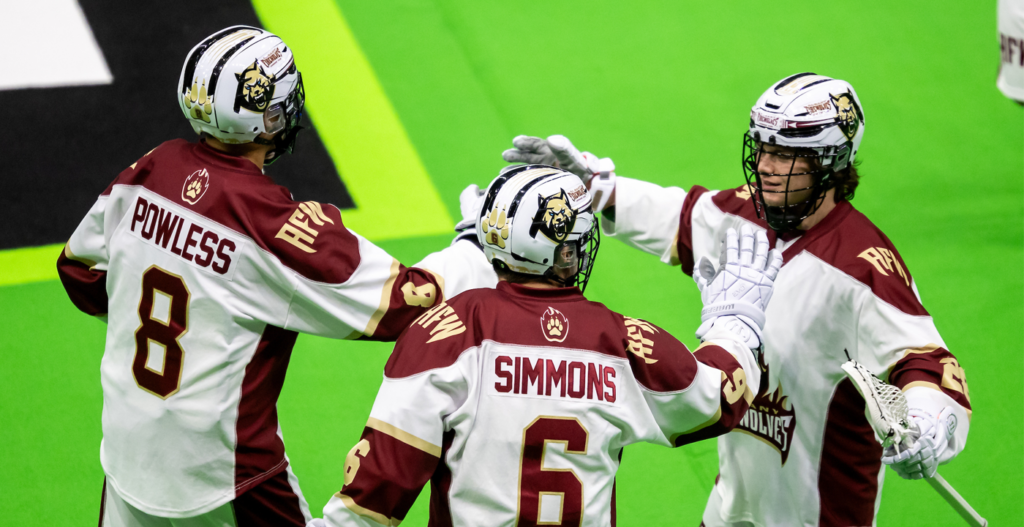 Three lacrosse players wearing team jerseys and helmets are celebrating on a bright green field. The player on the left has "POWLESS" and the number 8 on their jersey, while the center player has "SIMMONS" and the number 6. The third player, on the right, appears to be congratulating the others.