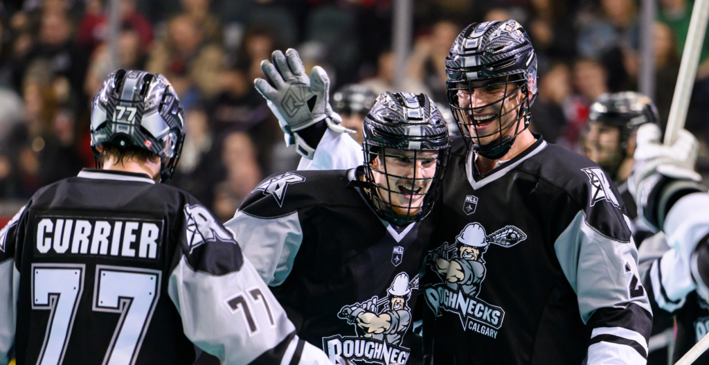 A group of lacrosse players in black uniforms celebrate a score, with one player raising his hand in excitement. The back of one player's jersey shows the name "CURRIER" and the number 77.