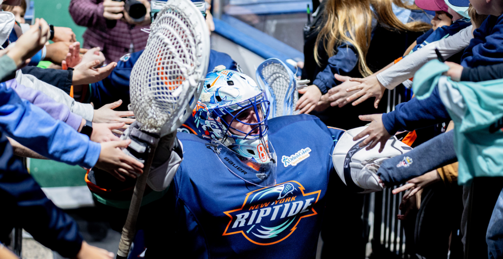 A lacrosse goalie in a blue jersey with "New York Riptide" on it interacts with fans, reaching out as they extend their hands in excitement. The goalie is wearing a helmet and holding a lacrosse stick, with numerous fans reaching out to touch him.