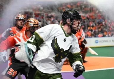 A lacrosse player in a white and green jersey with a bird logo, wearing a helmet and gloves, runs intensely on the field, while two opposing players in orange jerseys closely pursue him in the background. The stadium is filled with fans.