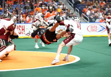 A lacrosse player performs a diving shot while being defended during a game, with spectators in the background.