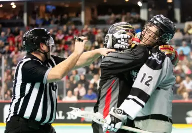 Referee intervenes as two lacrosse players grapple on the field during a game.