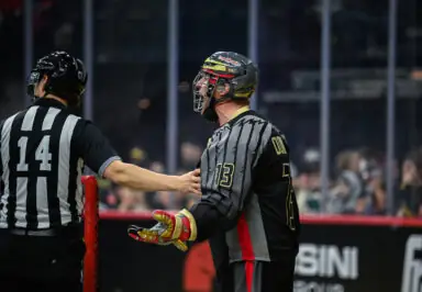 A lacrosse player in a gray and black jersey with the number 13 gestures emphatically while facing a referee, who is wearing a black and white striped uniform and has his hand on the player