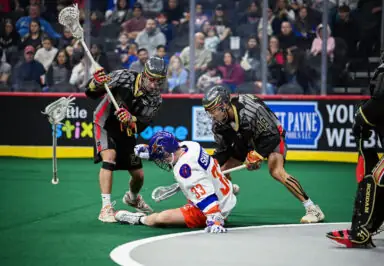 Two lacrosse players in black jerseys are attempting to gain possession of a ball from a player in a white and orange uniform who is on the ground. The audience is visible in the background.
