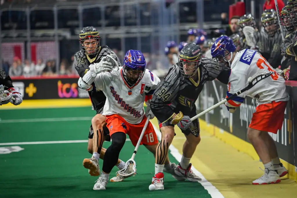A lacrosse game scene featuring players in action, with two players in white jerseys (one wearing number 18) battling for the ball against three players in dark jerseys (one wearing number 94) near the edge of the field. The setting includes spectators in the background and a green turf surface.