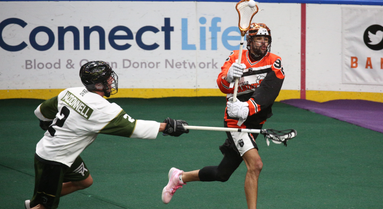A lacrosse player in an orange and black uniform dodges a defender wearing a green and white uniform on the field.