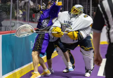 A lacrosse player in a purple jersey is being pushed against the glass by a goalie in white gear, with a stick involved in the action.