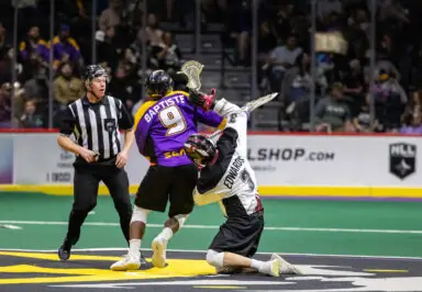 A lacrosse game scene showing a player in a purple jersey with the name "Baptiste" facing off against another player in a white jersey, while a referee watches nearby. The game is taking place on a green turf field with an audience in the background.