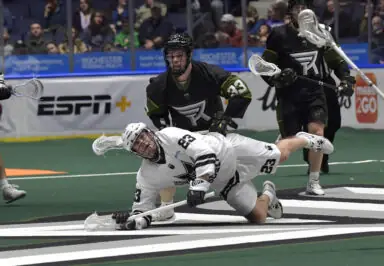 A player in a white uniform dives on the field while being pursued by two players in black uniforms during a National Lacrosse League game.