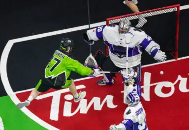 A lacrosse player in a bright green uniform, number 17, is in mid-air attempting a shot on goal while a goalie in a white and purple uniform defends the net. The floor is marked with a red-and-white logo, and another player in a white uniform stands nearby.