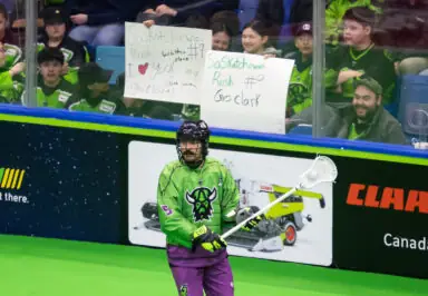 A lacrosse player in a green jersey stands on the field, with fans in the background holding signs in support.