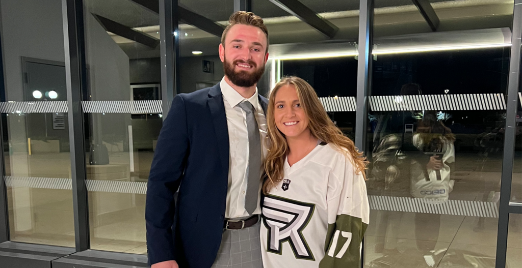 A man in a suit stands beside a woman wearing a National Lacrosse League jersey, both smiling in front of glass doors at night.