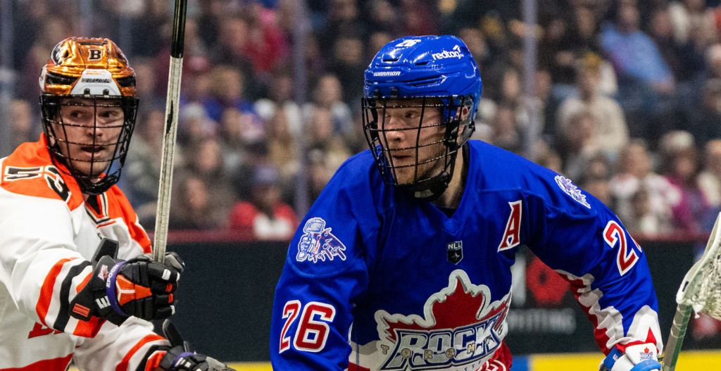Two lacrosse players engage on the field. One wears a blue jersey with "Toronto Rock" logo and the number 26, while the other is in an orange and white jersey, holding a stick. The crowd is visible in the background, indicating a lively event.