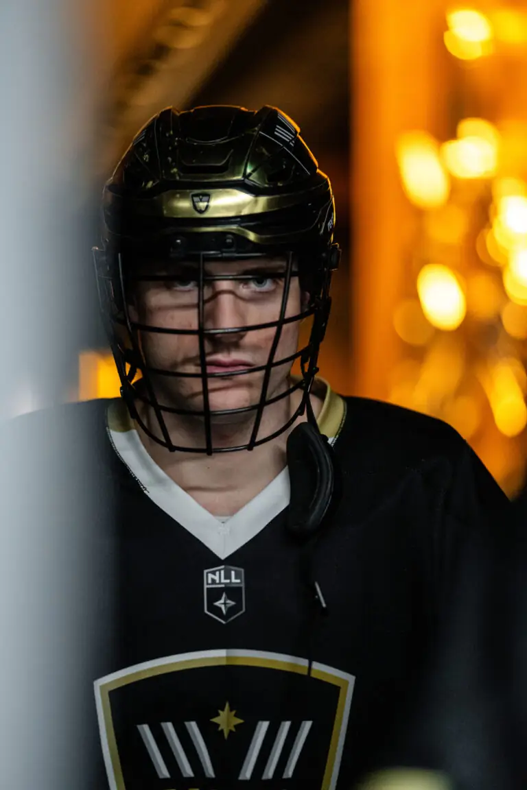 Lacrosse player wearing a helmet and jersey, standing in a dimly lit arena.