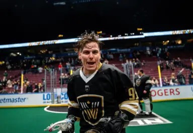Lacrosse player in a black jersey smiles and holds a stick on the field, with fans in the background.