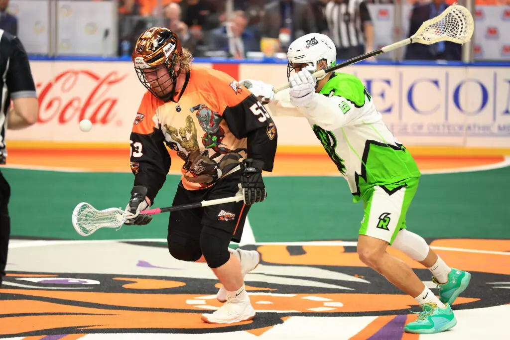 Two lacrosse players compete for a loose ball on the field. One player wears an orange jersey with a graphic design, while the other is in a bright green and white uniform.