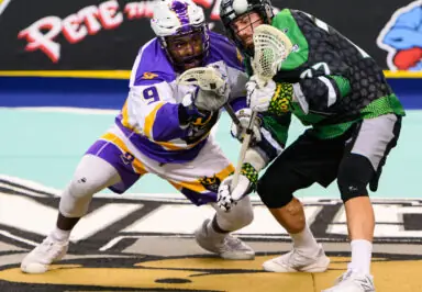 A lacrosse player in a purple and gold uniform fights for possession against an opponent in a green and black uniform during a National Lacrosse League game.