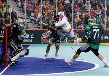 A lacrosse player leaps mid-air while attempting a shot, surrounded by defenders and a goalie in an indoor arena filled with spectators.