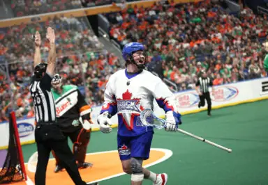 A lacrosse player in a white and blue jersey celebrates on the field as a referee signals a goal, with a crowd in the background.
