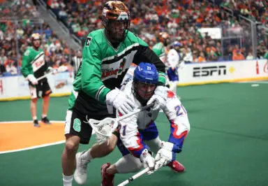 A player in a green jersey grapples with a player in a white jersey on the lacrosse field during a game.
