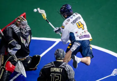 A lacrosse player in a white jersey attempts a shot on goal while a goalie in black defends, with another player in black nearby.