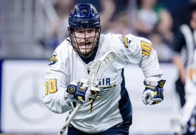 A lacrosse player in a white and blue uniform runs with a stick, showcasing intense focus during a game.