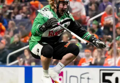 Lacrosse player in a green and black Buffalo Bandits uniform jumping with a stick in hand, during a game with a cheering crowd in the background.