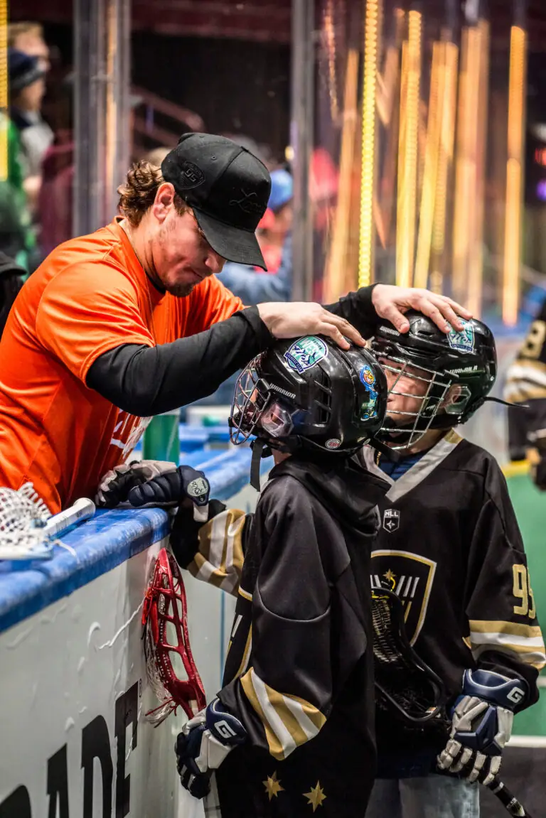 A player in an orange shirt interacts with two young lacrosse players in helmets and jerseys at the rink.