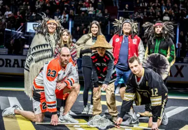 Participants in lacrosse uniforms and traditional attire pose together on the field.