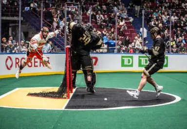 A player in a white jersey leaps towards the goal as a goalie in black prepares to block a shot during a National Lacrosse League game.
