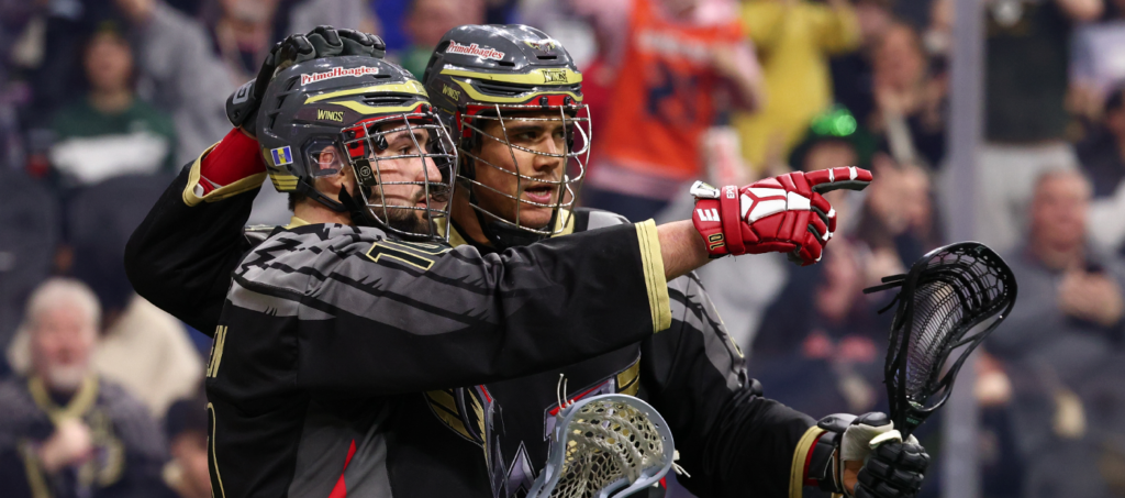 Two lacrosse players celebrate together on the field, one pointing while the other smiles, wearing team uniforms and helmets.