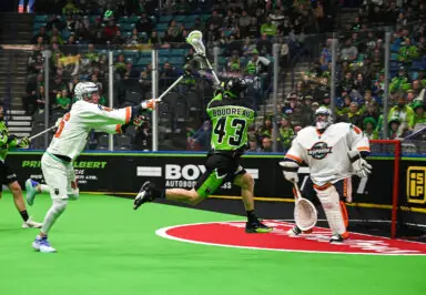 A player in a green lacrosse uniform shoots the ball while another player in white defends, with a goalie in the background.