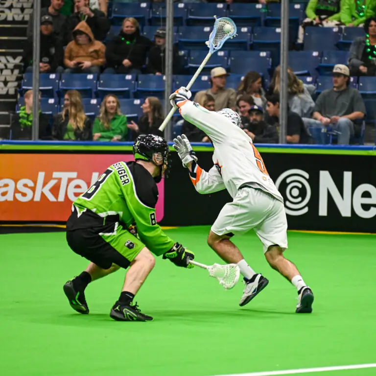 A player in a white lacrosse uniform is about to pass the ball while another player in a green uniform attempts to check him. Fans are visible in the background.