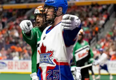 A lacrosse player in a white and blue jersey with a maple leaf design celebrates while pointing, with another player in green behind him.
