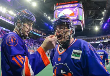 Two lacrosse players in blue jerseys discuss on the sidelines, one adjusting the other’s mouthguard, with a crowd and large screen in the background.