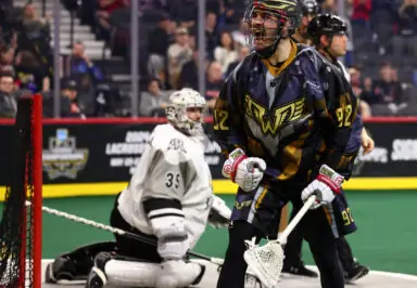 A lacrosse player celebrating near the goal, with a focused goalie in the background.